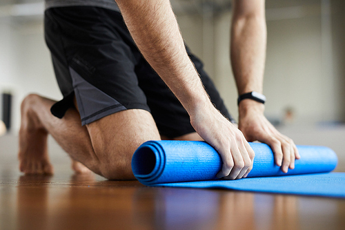 Close-up of unrecognizable man standing on knees and rolling out exercise mat after yoga training in gym