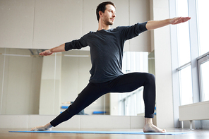Content slim handsome young man in leggings keeping one foot forward and outstretching arms while doing standing yoga pose