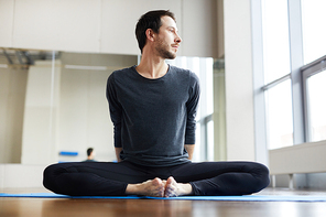 Serious handsome flexible young man with stubble sitting inbound angle pose on yoga mat and looking aside while stretching in studio