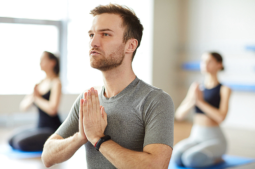 Serious peaceful handsome young man with stubble concentrated on thoughts joining hands in Namaste and meditating at yoga class