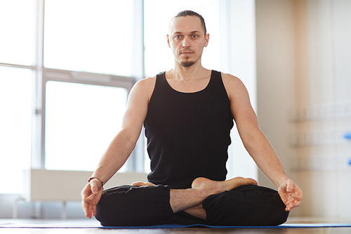 Serious muscled young man with beard sitting in lotus position and meditating alone in empty room, he 
