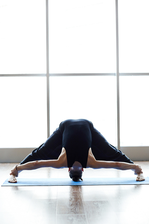 Flexible man standing on exercise mat against window and doing wide-legged standing forward bend