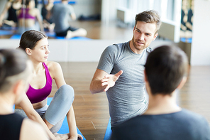 Serious pensive sporty guy with stubble sitting on floor and gesturing hand while sharing ideas for good training at yoga class