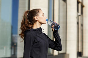 Serious thirsty attractive female jogger with ponytail wearing track suit standing against city building and drinking water from bottle