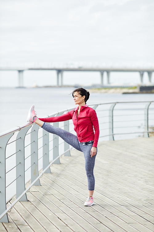 Full length portrait of active mature woman stretching legs outdoors while jogging on pier, copy space