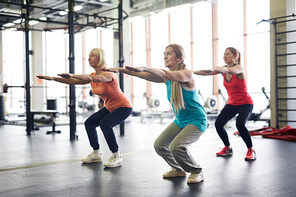Group of mature active females squatting and stretching arms forwards during workout in fitness center
