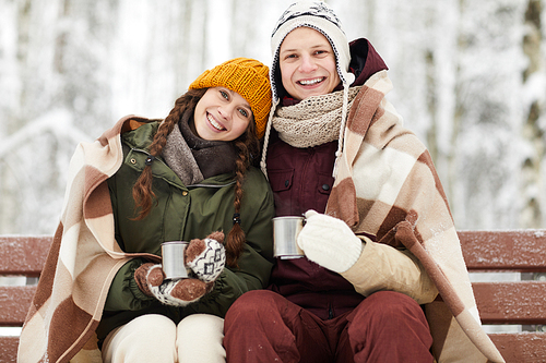 Waist up portrait of loving young couple wrapped in blanket drinking hot cocoa outdoors while sitting on bench beautiful winter forest, copy space