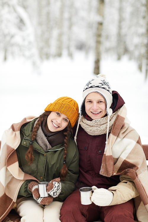 Portrait of loving young couple wrapped in blanket drinking hot cocoa outdoors while sitting on bench beautiful winter forest, copy space