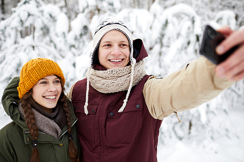 Waist up portrait of happy young couple taking selfie outdoors in beautiful winter forest