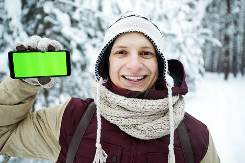 Portrait of active young man showing smartphone with green screen during hike in winter forest