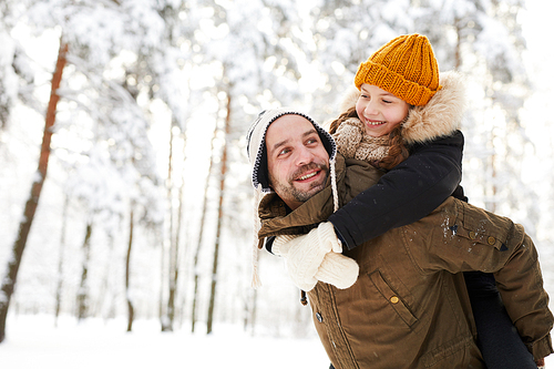 Portrait of happy little girl riding on fathers back while having fun in winter forest, copy space