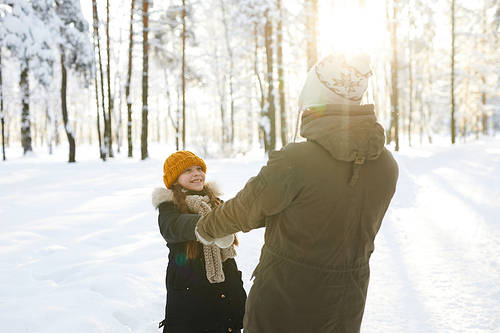 Portrait of happy little girl holding hands with dad while having fun in winter forest lit by sunlight, copy space