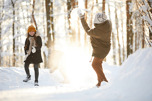 Full length portrait of happy little girl throwing snow at father while having fun in winter forest lit by sunlight, copy space