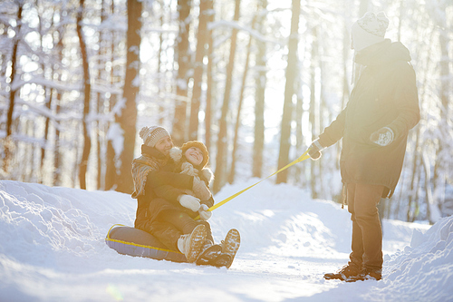 Full length portrait of happy family enjoying sleigh ride in winter forest lit by sunlight, copy space