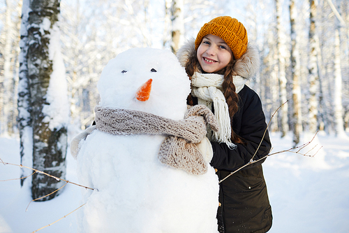 Portrait of happy little girl building snowman in winter forest and smiling at camera, copy space
