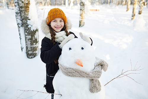 Portrait of happy girl building snowman in winter forest and smiling at camera, copy space