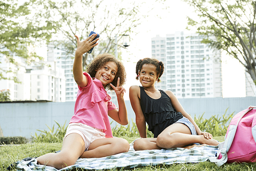 Jolly excited teen black girls in casual clothing sitting on blanket in city park and using smartphone while posing for selfie together