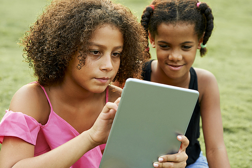Serious curious African girls sitting on grass in park and using digital tablet while surfing net together