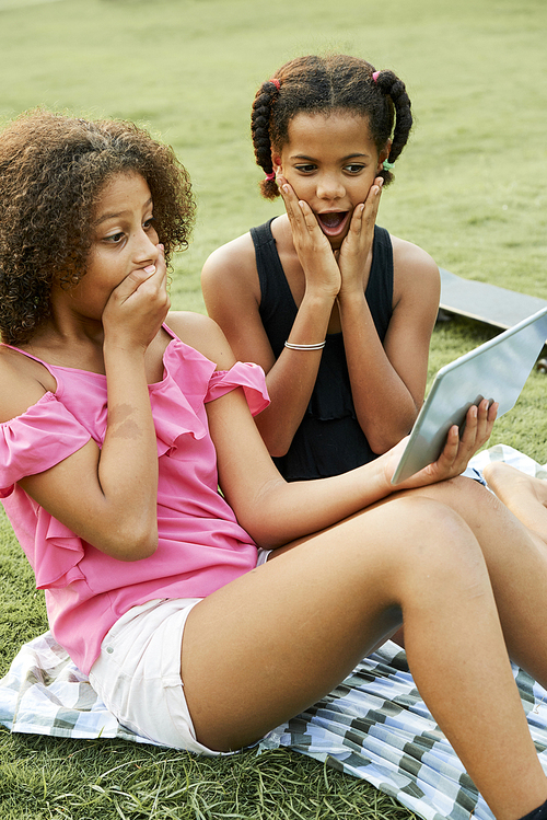 Surprised excited teen black girls sitting on ground and using digital tablet while watching video online, one girl covering mouth with hand while her friend keeping mouth open