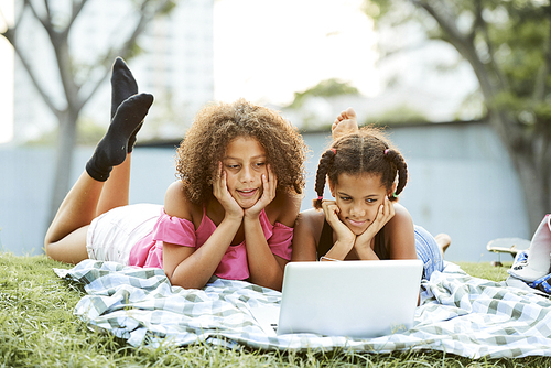 Content curious African-American girls in casual outfits lying on ground and leaning heads on hands while watching interesting video on laptop