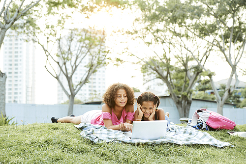 Positive excited African-American school friends lying on grass in public park and using modern laptop while enjoying movie at picnic