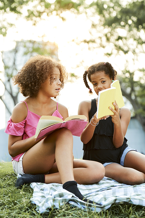 Positive curious African girls sitting on blanket in city parks and reading books while sharing useful information