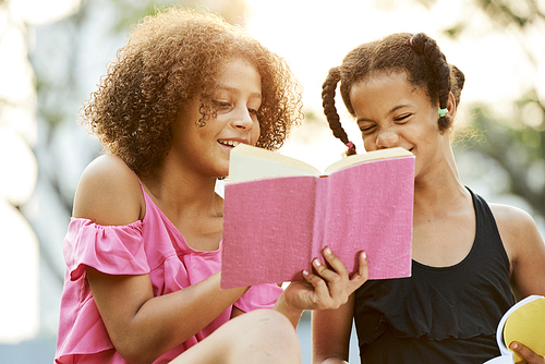 Cheerful excited teenage black girls with Afro hairstyles sitting in park and laughing while reading interesting book together
