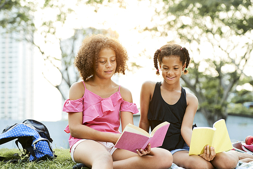 Cheerful curious African girls in casual clothing sitting on grass in park and reading books while getting knowledge during outdoor picnic