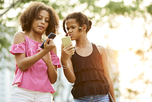 Confused African-American girls in casual clothing standing outdoors and checking information on smartphones
