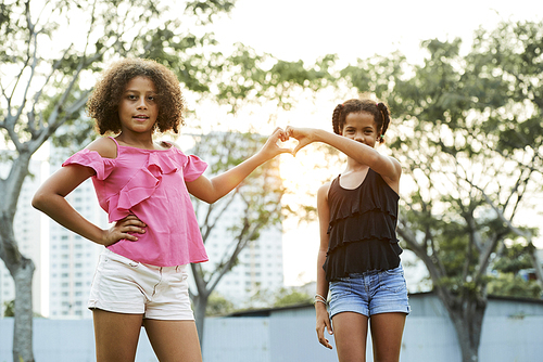Cheerful positive African girls in casual clothing standing against cityscape and joining hands in shape of heart while 