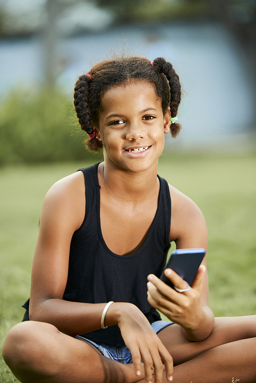 Smiling attractive Black girl with funny braids sitting with crossed legs on grass outdoors and  while using modern smartphone