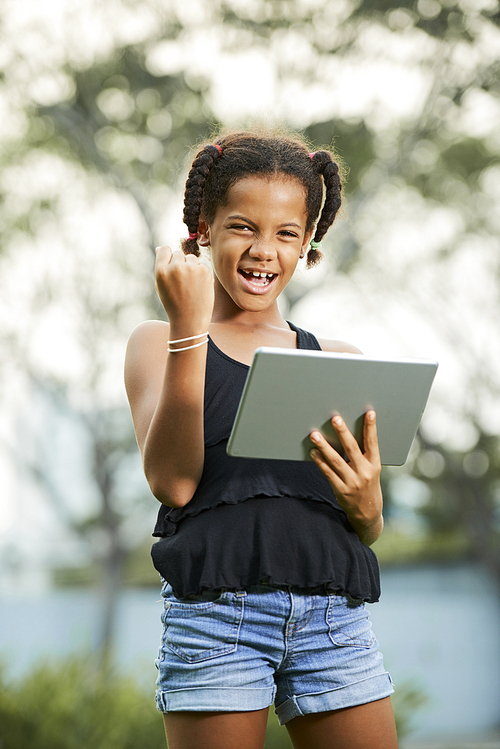 Cheerful successful African-American teen girl with braids standing in park and making yes gesture while using digital tablet