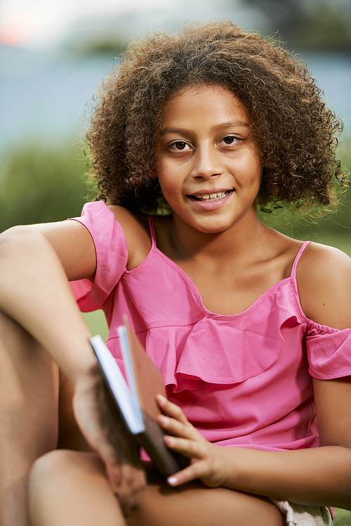 Positive pretty teenage black girl with Afro hairstyle wearing stylish blouse , she reading book in park