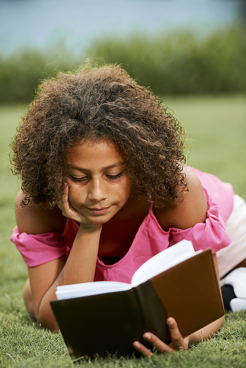 Serious concentrated curious African girl with curly hair lying on grass in park and leaning head on hand while reading literature