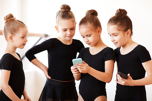 Group of little girls using smartphone standing in circle during break in ballet class, copy space