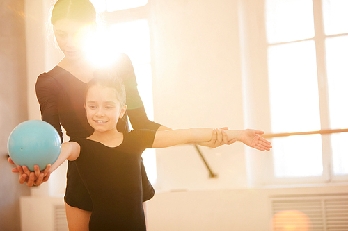 Portrait of  woman teaching little girl doing gymnastics moves with ball in studio lit by warm sunlight, copy space