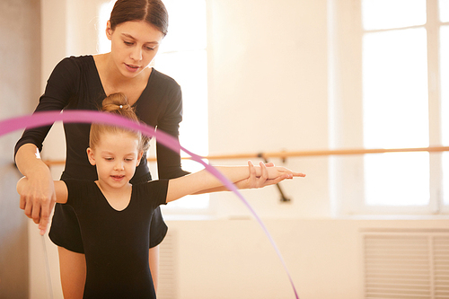 Portrait of woman teaching little girl doing gymnastics moves with ribbon in studio lit by warm sunlight, copy space