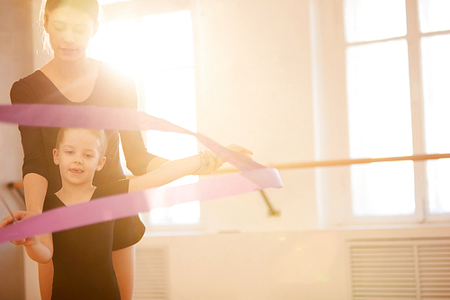 Portrait of coach teaching little girl doing gymnastics moves with ribbon in studio lit by warm sunlight, copy space