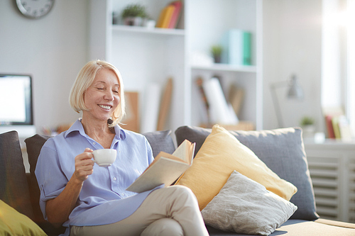 Portrait of contemporary adult woman reading book sitting on sofa at home and smiling happily, copy space