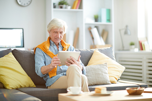 Portrait of contemporary senior using digital tablet sitting on sofa at home, copy space