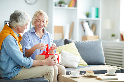 Portrait of happy mature couple exchanging gifts at home focus on woman unwrapping gift box, copy space