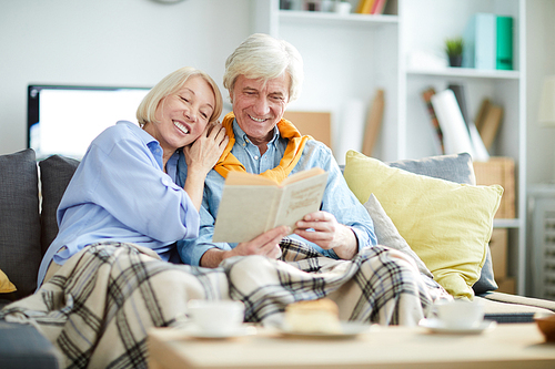 Portrait of loving mature couple reading book together sitting on sofa at home, copy space