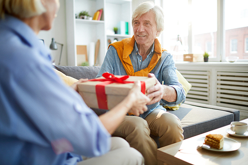 Portrait of contemporary mature couple exchanging gifts at home, focus on man holding present, copy space