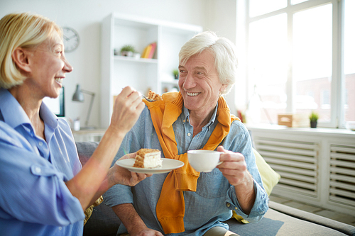 Portrait of happy mature couple enjoying tea and cake at home lit by sunlight, copy space