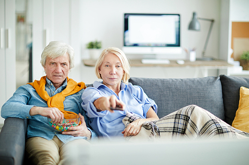 Portrait of contemporary mature couple watching television together at home sitting on comfortable sofa, copy space