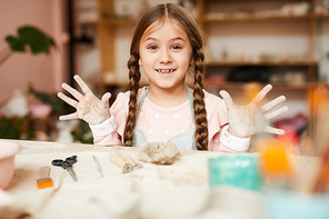 Portrait of cute little girl posing in pottery class showing hands smudged with clay, copy space