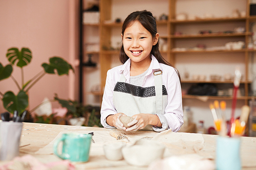 Portrait of Asian girl smiling at camera while enjoying pottery class in workshop, copy space