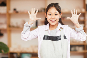 Waist up portrait of happy Asian girl posing in pottery workshop showing hands covered with clay to camera, copy space