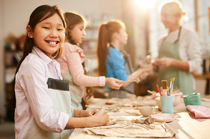 Waist up portrait of happy Asian girl looking at camera while enjoying pottery class with group of children, copy space