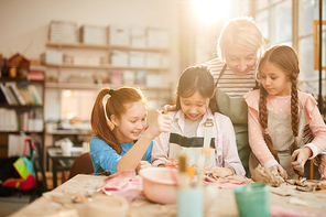 Portrait of female art teacher working with little girls in pottery class, scene lit by serene sunlight, copy space